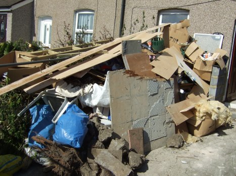 A pile of mixed waste and building debris is accumulated on a sidewalk area, consisting of broken bricks, concrete blocks, and fragments of plaster, alongside discarded cardboard boxes, some flattened and others partially intact. The debris includes pieces of wood, such as a long, narrow wooden plank leaning diagonally across the heap, and torn, crumpled fabric or insulation material amongst the rubbish. A blue plastic bag is visible near the bottom left, partially buried under the rubble. The waste is situated against the exterior wall of a residential building with textured, grey stucco finish and two white-framed windows with closed curtains. In the background, neighboring houses with similar architectural features can be seen, indicating a residential street environment. The scene appears to be during daylight hours, with natural sunlight casting clarity over the accumulated waste and surroundings. This image showcases typical clutter requiring professional rubbish removal, as provided by Brixton House Clearance in the London area, demonstrating the type of debris handled during house clearance or renovation waste disposal in Brixton or nearby postcode districts.
